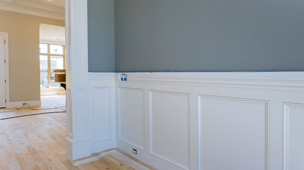 A painting project in progress or an interior wall with waist height white trim board and gray paint, to left is a larger room leading to doors and windows, with lighter colored hard wood floors.